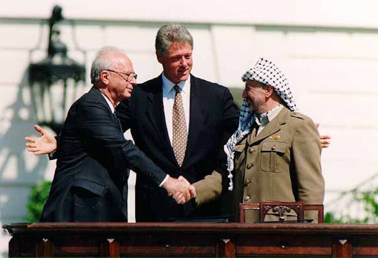 Isaac Rabin y Yasser Arafat se dan la mano mientras Bill Clinton abre los brazos detrás de ellos.