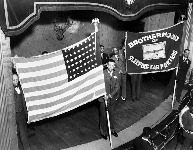 Black men stand on a stage holding an American flag and a union flag.