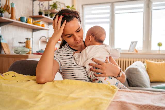 Young adult mother sitting in chair at home, holding her 3-month old baby son, with a tired expression on her face.