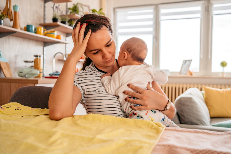 Young adult mother sitting in chair at home, holding her 3-month old baby son, with a tired expression on her face.