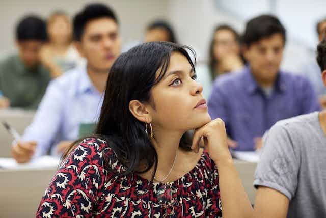 A female student with her chin resting on her hand sits at a desk in a college classroom apparently looking at her instructor.