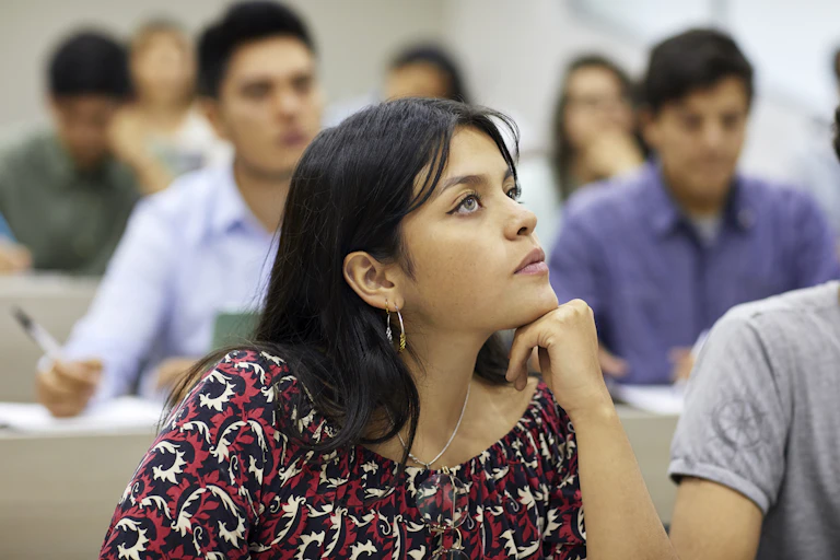 A female student with her chin resting on her hand sits at a desk in a college classroom apparently looking at her instructor.