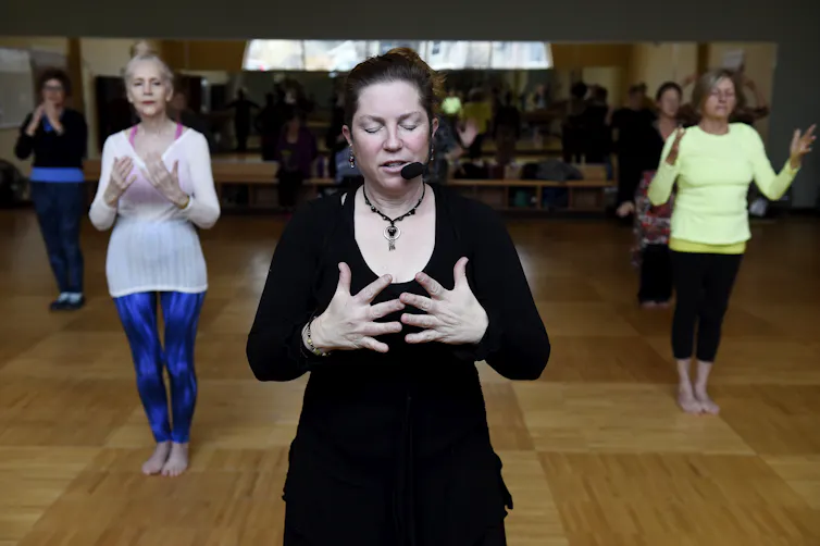 A woman wearing a headset microphone leads a class of women, all holding their palms in front of their chests. The instructor has her eyes closed.
