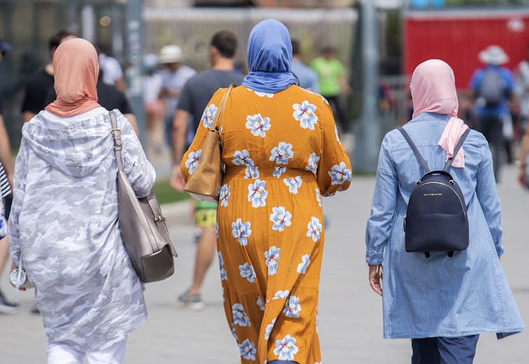 Three women wearing hijabs are seen walking along a street. One carries a knapsack on her back.