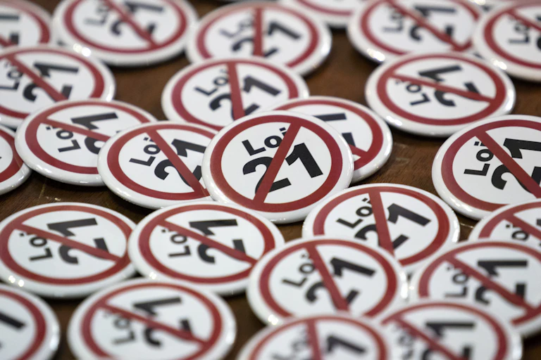 A collection of red, black and white lapel pins that say Loi 21 with a red slash through it.