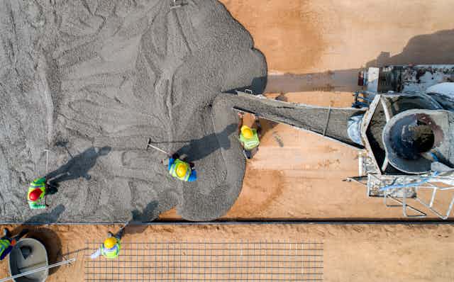 A machine pours cement for concrete while workers in hardhats smooth it out. 