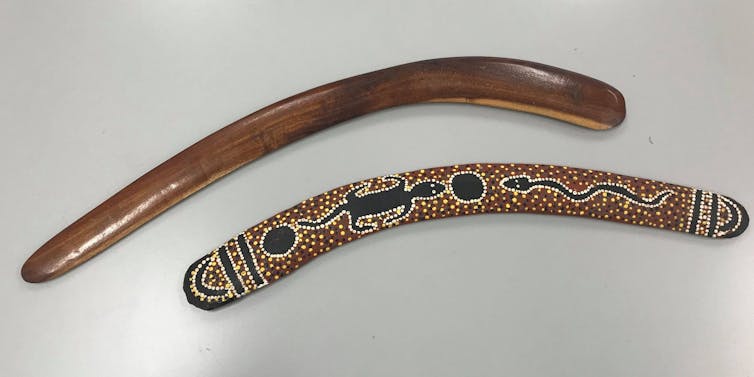 Two brown, slightly curved'sticks' on a white surface, one decorated with an Indigenous art style