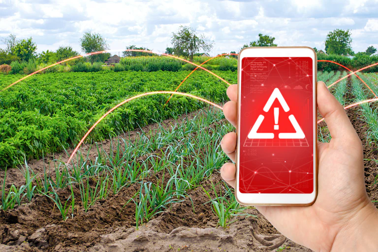A hand holds a cellphone whose screen contains a white triangle and exclamation mark against a red backdrop. There are crops and trees in the background of the image