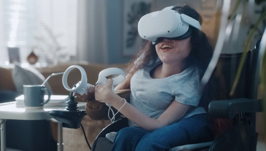 A woman in a wheelchair smiles while wearing a VR headset and operating two controllers with her hands