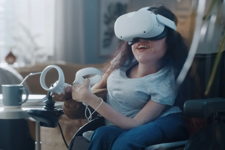 A woman in a wheelchair smiles while wearing a VR headset and operating two controllers with her hands