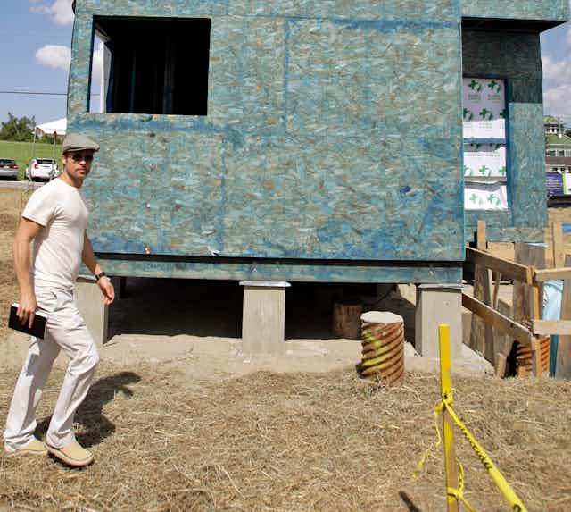 Man dressed in white wearing a cap walks past a house under construction.