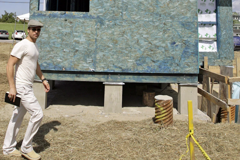 Man dressed in white wearing a cap walks past a house under construction.