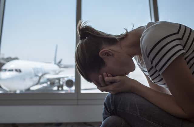 A despondent young woman, her head covered by her hands, sits forlornly inside an airport terminal.