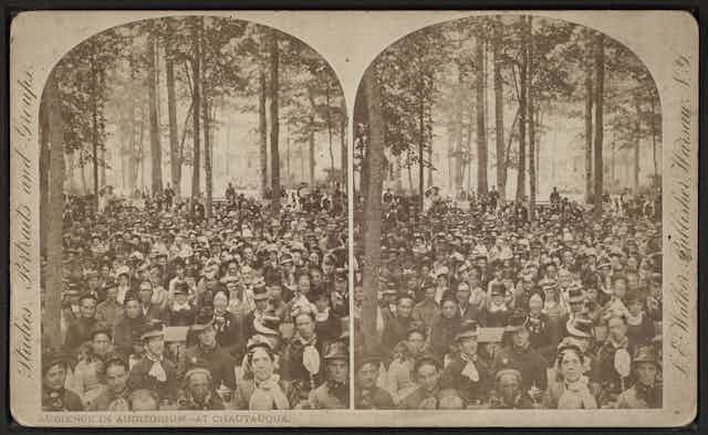 A black and white postcard shows two copies of a photograph of a large crowd seated among trees.