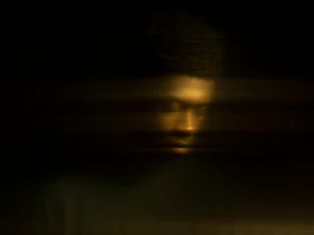 a dark background and a man's face, blurry, looking down
