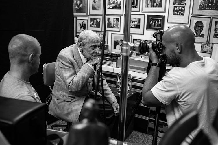 An elderly man leans forward in a chair as a photographer takes his photo.