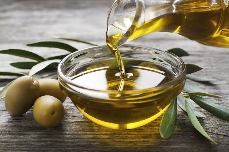 Close up photo of olive oil being poured from a glass pitcher into a glass bowl, with olives and leaves on the table surrounding