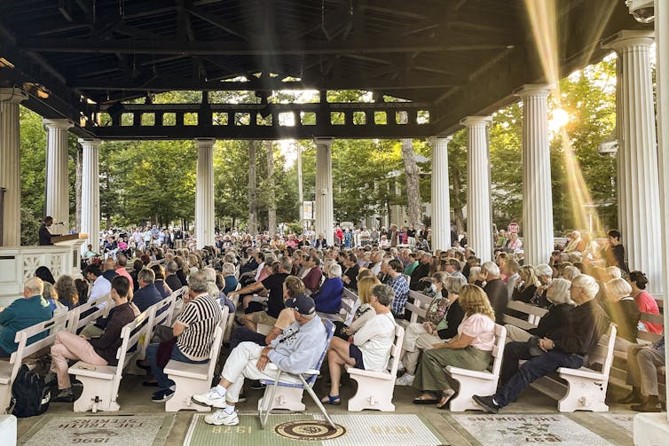 Half a dozen rows of people sit underneath an outdoor shelter among trees.