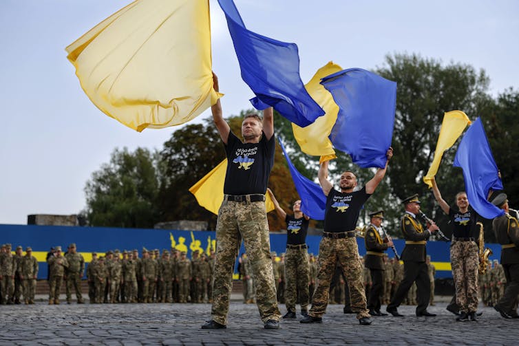 Men and women wearing camouflage pants and black T-shirts line up and raise blue and yellow flags above their heads.