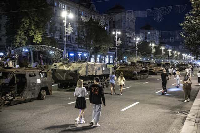 A dark city street has military tanks, some looking destroyed, lining the empty avenue. Young people are seen walking casually down the street.