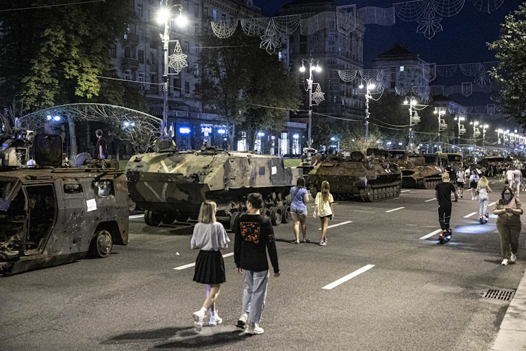 A dark city street has military tanks, some looking destroyed, lining the empty avenue. Young people are seen walking casually down the street.