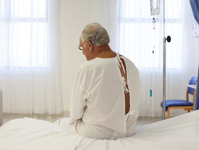 A man with white hair, wearing a hospital gown, seen from behind sitting on a bed
