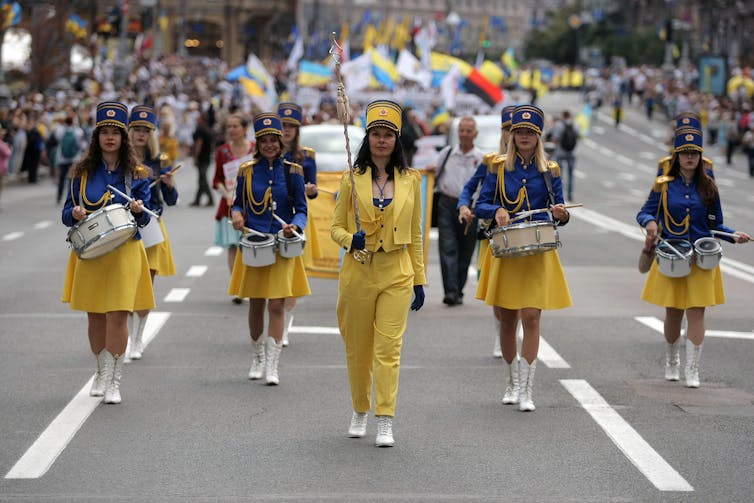 Women in yellow skirts and blue jackets and hats smile while holding small drums during the parade, while a woman in yellow pantsuits leads them with sticks.  A large parade of people carrying flags stands behind them.