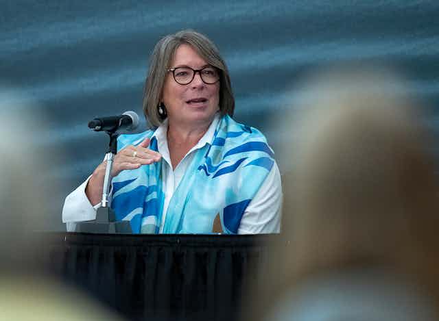 A woman wearing a white top and blue shall stands behind a microphone