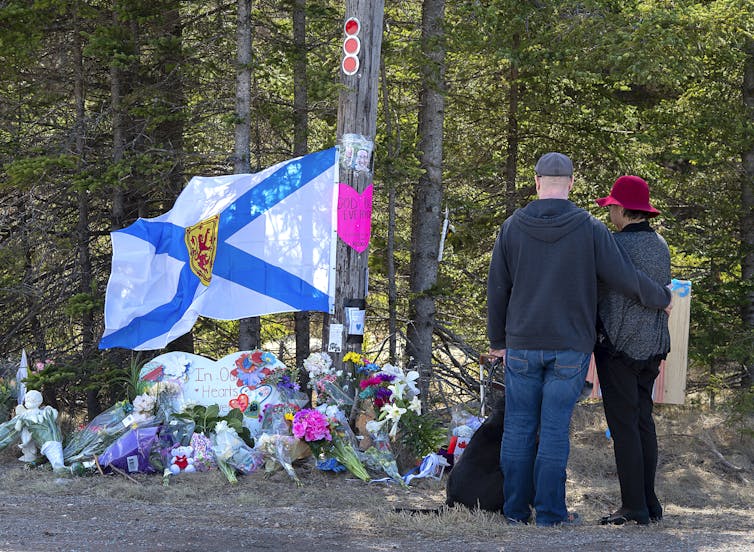 A Nova Scotia flag hangs above flowers at a makeshift memorial. A couple stands looking onward