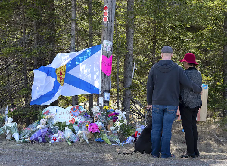 A Nova Scotia flag hangs above flowers at a makeshift memorial. A couple stands looking onward