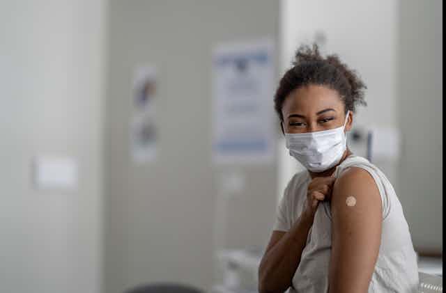 A young Black woman, in a clinic and wearing a mask, shows off the Band-Aid that covers her vaccine mark.