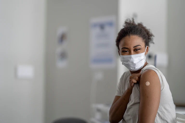 A young Black woman, in a clinic and wearing a mask, shows off the Band-Aid that covers her vaccine mark.