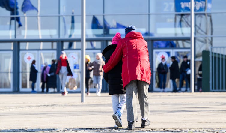 A tall person in a red coat accompanies a smaller person toward a glass building.