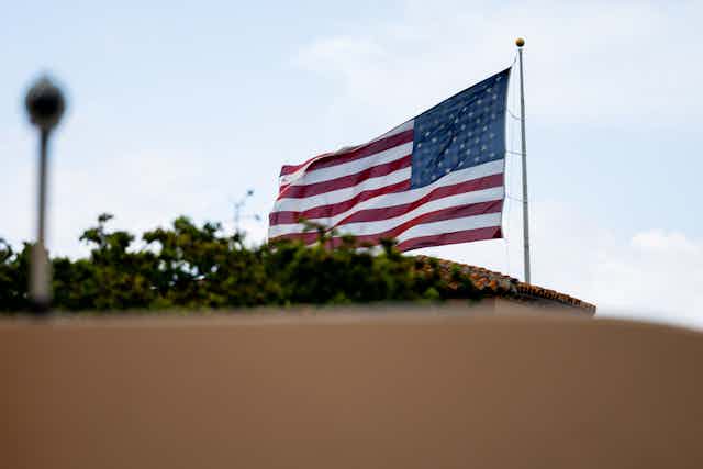 An American flag is shown waving above a brown barrier and above tree tops.