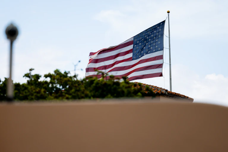 An American flag is shown waving above a brown barrier and above tree tops.