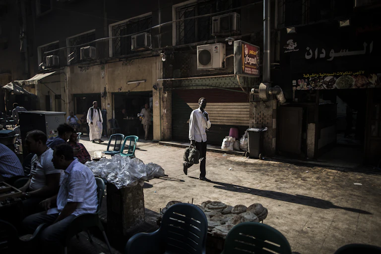 A man walks down a street with a Sudanese flags and men sitting at tables