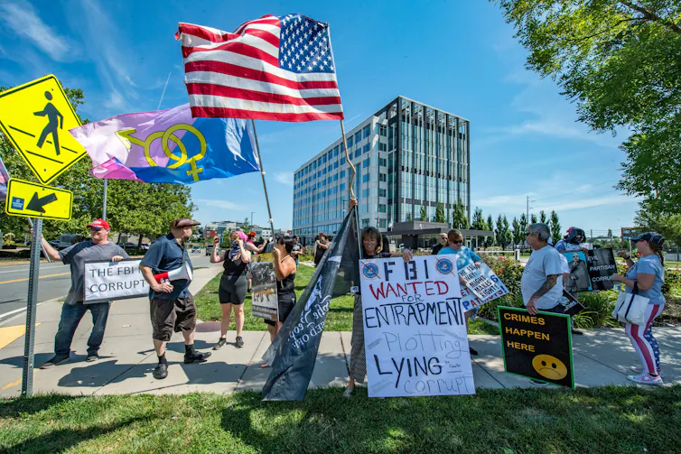 People hold flags and signs that say 'The FBI is corrupt' and 'crimes happen here' on a street corner on a sunny day.
