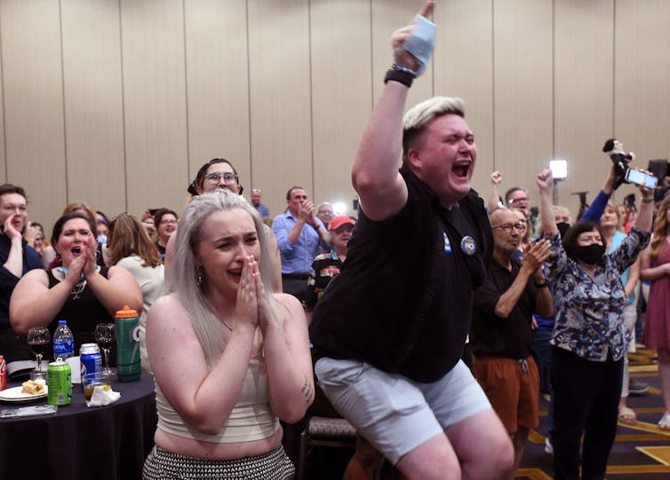 A room of people are seen jumping, crying, cheering and raising their arms. Int he front are two young people who appear emotional and happy.