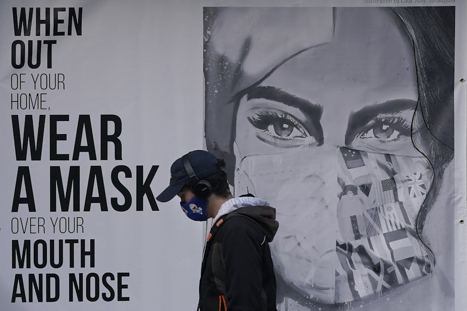 Person wearing mask walking in front of poster of mural reading "When out of your home, Wear a mask over your mouth and nose"