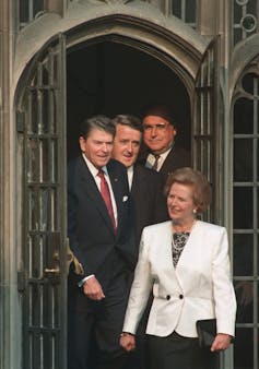 Three men follow a woman in a white suit jacket out of a building. All are smiling.