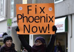 A woman in a hat and mitten carries an orange sign that reads Fix Phoenix Now.