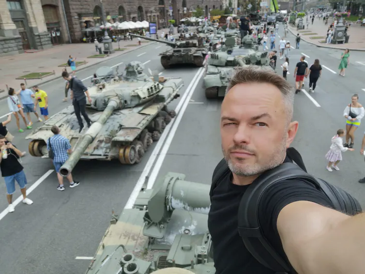 A man takes a selfie with destroyed military vehicles in the background.