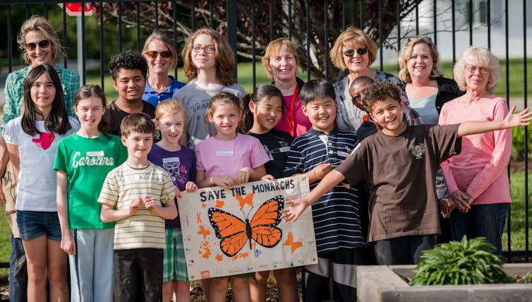 A group of adults and children hold a banner that reads 'Save The Monarchs'.