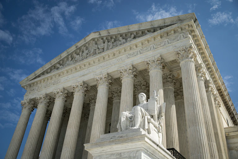 A view of the front of the US Supreme Court building with columns and a statue of a seated person.