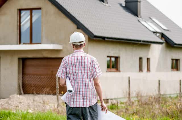 Man wearing hard hat with rolled-up blueprints under his arm stands in front of a house