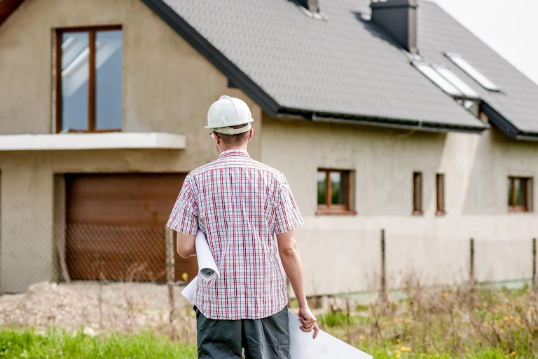 Man wearing hard hat with rolled-up blueprints under his arm stands in front of a house