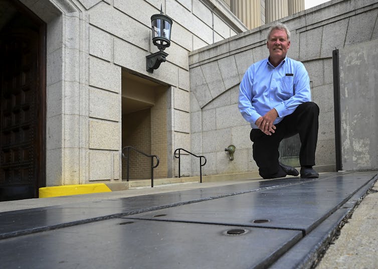 A man stands next to a long metal door that opens from the ground and is part of a flood control system for the building behind him.