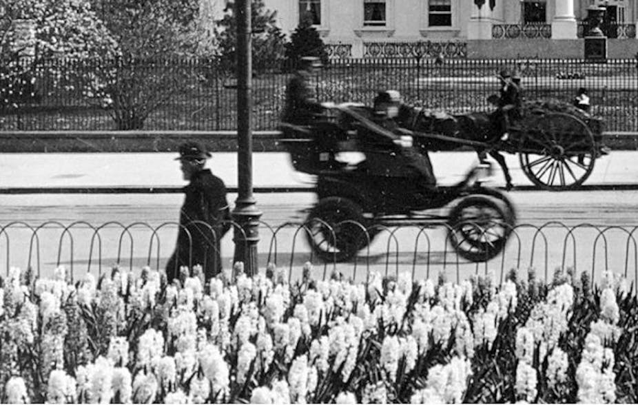 Very early 20th-century electric cab drives past the White House in front of a horse-drawn carriage