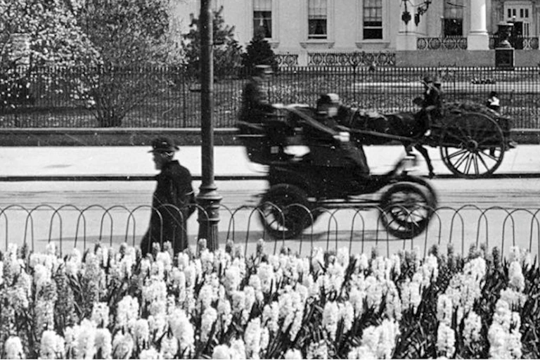 Very early 20th-century electric cab drives past the White House in front of a horse-drawn carriage