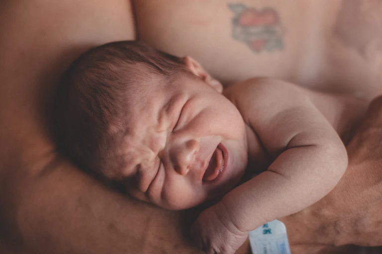 A baby is cradled by a shirtless person with tattoo on chest.